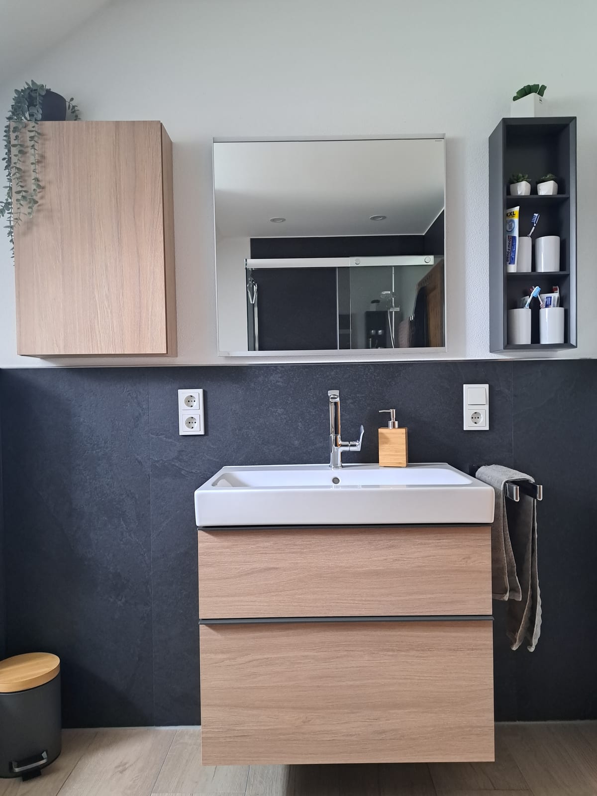 Waschtisch mit Holzfront und goldfarbenen Accessoires vor anthrazitfarbener Fliesenwand