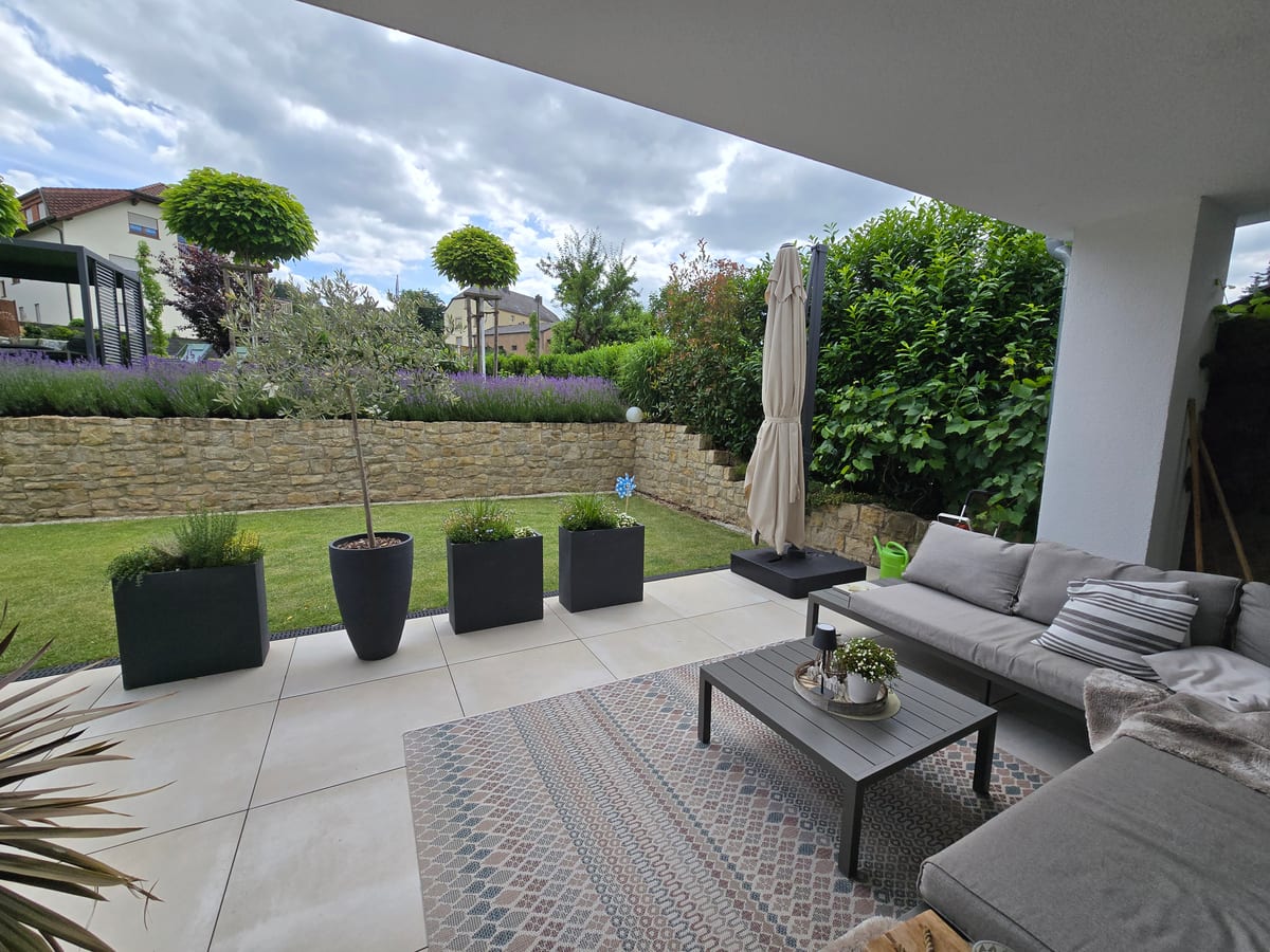 Terrasse mit großformatigen hellgrauen Bodenplatten, Loungemöbeln und gepflegtem Garten in Luxemburg